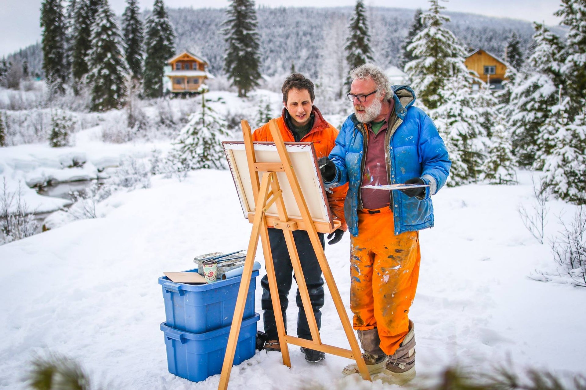 Load video: A video of Peter Corbett painting outdoors by a creek side with television host. Background scenery is snowy-laden trees and cabins off in the distance. In the foreground is an easel and painting supplies